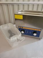 Ultrasonic cleaner with the metal cleaning basket wrapped in bubble wrap placed beside it, showing the front control panel and stainless steel tank.