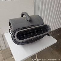 Photo showing front and side view of gray air mover fan with oval air outlet and carry handle, power cord coiled on side.