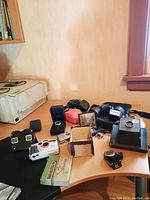 Photo showing various items on wooden table including cameras, binoculars, meter, watch, and View Master viewer.