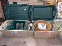 Two large Rubbermaid Rough Tote rectangular plastic storage bins with snapped-on dark green lids open, showing contents including green plastic laundry basket and wooden bushel basket with white plastic container inside.