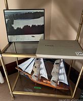 Shelf view showing the framed photograph of a snowy landscape with a horse and sled, the hardboard sleeve of the coffee table book 'Canada: A Year of the Land', and the wooden model ship with plastic sails beneath it.