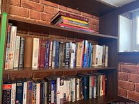 Overview of bookshelves containing a variety of hardcover and paperback books including novels, history books, and magazines arranged on wooden shelves against brick background.