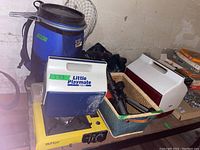 Wide shot of the camping lot items on a table including Burton burner, coolers, fishing net, basket with gear, and blue water jug backpack