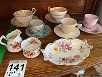 Overview photo showing multiple porcelain cups, saucers, a creamer, sugar bowl, and a floral Royal Crown Derby trinket dish with gold detailing on a wooden surface.