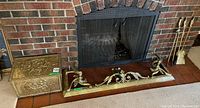 Photo showing ornate brass firewood box with embossed floral motif and brass fire guard with intricate scrolling design in front of a brick fireplace.