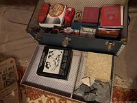 Photo showing open blue metal case containing multiple assorted vintage game boxes and books, some with visible titles, and several fossil rocks in the lower compartment
