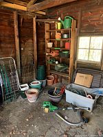 Photo showing various clay and plastic planters, gardening tools on floor including John Deere gas trimmer, plant stakes and supports, and plastic pots on shelf in wooden shed with window light.