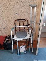 Wide view showing white and gray toilet seat with arm handles, brown-handle walker behind it, crutches leaning on wall, and small items on floor.