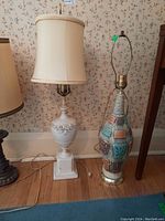 Two table lamps on a wooden floor by a floral wallpapered wall; left lamp has white porcelain base and fabric shade, right lamp has multicolored ceramic base without shade.