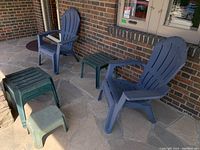 Two blue plastic Muskoka style patio chairs next to four green small slatted side tables on stone patio floor against brick wall