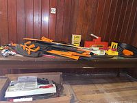 Overview of all race track pieces, cars, and train tracks on a table in front of a wood panel wall