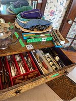 Oak drawer open showing organized silver plate cutlery and kitchen tools inside, along with a woven basket and fabric bags on top of a wooden furniture piece.