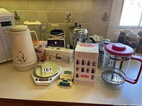 Wide view of assorted kitchen appliances and accessories on countertop including thermos, food scale, boxed juicer and blender, Bodum French press and small kitchen items.