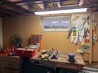 Wide view of basement workbench with toolbox, fan, small organizer cubbies, hardware and various hand tools laid out.