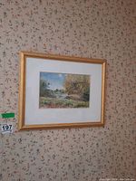 Framed watercolour landscape painting on wallpapered wall, showing gold-tone wood frame and white matting surrounding the artwork.