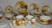 Overview of entire Noritake hand painted dinnerware set arranged together on a white cloth backdrop, showing plates, bowls, cups and condiment set with brown and beige scenic artwork.