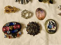 Top-down view of nine assorted vintage brooches with crystal accents and painted cameo styles on a white fabric surface.