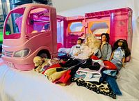 Pink Barbie Glamour RV motor home camper van fully set up with four dolls seated and several piles of doll clothes laid out alongside.