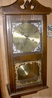 Front angled view of the Daniel Dakota pendulum wall clock showing wooden case, brass dial with Roman numerals, and pendulum behind decorative glass panel.
