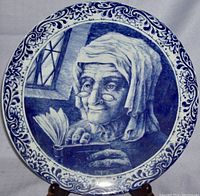 Front view of the circular ceramic charger plate showing an elderly woman in blue cobalt ink reading a book, framed by a detailed floral border.