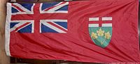 Photo showing the red Ontario flag with British Union Jack and provincial shield, nylon material with visible texture and UV coating, edges reinforced with white fabric for hanging.