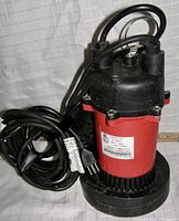 Front side view of the Red Lion RL-SP50T sump pump showing the red thermoplastic and black aluminum housing with power cord and plug coiled beside it.