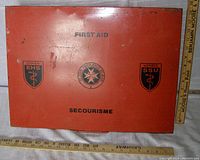 Closed red first aid metal case with medical logos and text ‘FIRST AID’ and ‘SECOURISME’. Shows external condition of case.