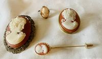 Overview photo of the four cameo jewelry pieces including two brooches, one ring, and one hat pin arranged on light fabric background.