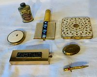 Top-down view of 7 vintage ladies accessories including compact powder cases, lipstick holder, talc tin, first aid kit and sword brooch