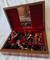 Overview of the open wooden jewelry box showing the red velvet interior, mirror, and assorted jewelry including rings, earrings, necklaces, and bracelets.
