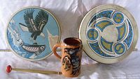 Front view of two West Coast style drums with native designs, a ceramic mug, and a drumstick laid out on a white cloth.