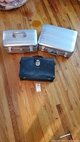 Two silver aluminum cases and one black case on a wooden floor. Aluminum cases rectangular with handles and locks, black case smaller with initials 'A.J.S.'