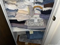 View of linen closet shelves with various folded towels, washcloths, and rugs in multiple colors including white, blue, beige, purple, and gray.