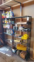 Full view of the black metal wire shelving unit standing against a wooden wall with various items stored on shelves (not included in lot).