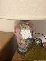 Close-up view of ceramic Chinese table lamp body showing detailed traditional artwork, lamp shade, and wood base with label attached.