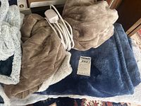Close-up photo showing a brown fleece heating blanket with controller on top of a navy blue heating blanket folded underneath, with a small MaxSold tag nearby.