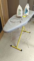 Full view of standing ironing board with two irons resting on it, cords visible