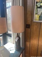 Full side view of mid century lamp on a blue tiled surface next to a window, showing ceramic striped base and tan fabric shade.