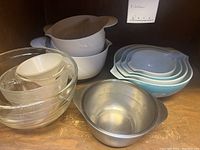 Photo showing variety of mixing bowls in clear glass, white ceramic, nested blue and white ceramic bowls, and metal bowl with handle and spout arranged in a wooden cabinet.
