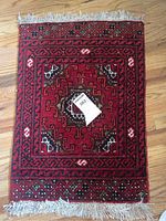 Full view of small red geometric patterned rug with fringed edges, placed on wooden floor with a tag labeled 'maxsold 302'.