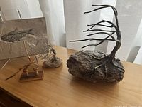 Photo showing three blowing tree sculptures placed on a wooden table: two metal trees on stone bases and one small wooden blowing tree sculpture. Also visible is the fossil fish stone slab on an acrylic stand.