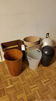 Photo shows six trash cans of different materials and colors on hardwood floor near wall.