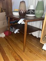 Side angle view of wooden side table showing tapered leg and lower shelf with hardwood floor background.