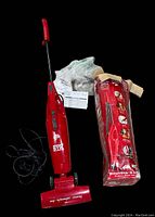 Red Dirt Devil SimpliStik bagless upright vacuum cleaner alongside original box and plastic wrapping, showing front and side views with power cord and control switch.