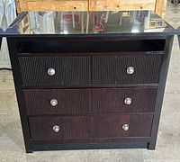 Front view of dark wood dresser with three drawers, each having two round metallic knobs. Smooth black marble top and open compartment above the drawers. No back panel visible.