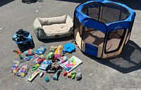 Full view of the lot items including blue pet playpen, beige pet bed, multiple cat toys, and blue pet carrier bag spread out on pavement