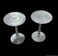 Two round steel stands side by side on a black background, showing top and base surfaces with worn metal finish.