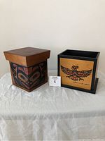 Two wooden First Nations Haida boxes side by side on a white draped surface against a plain wall. One is hexagonal with dark tones and red and black faces painted. The other is square with a bird design in black and red, signed.
