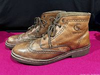 Side view of both distressed brown leather CANGURO men's boots on a pink surface, showing brogue style perforations and laces.