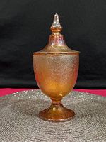 Full view of the 1920s Jeanette Marigold Carnival Glass Apothecary Jar with lid on a gray textured mat with black background.